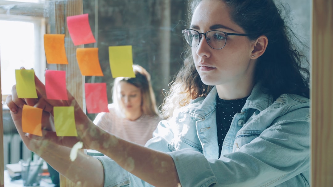 Close-up shot of attracvive young businesswoman standing and sticking colourful memos on glass board. Her colleague is sitting and working in background. Creative work concept.
